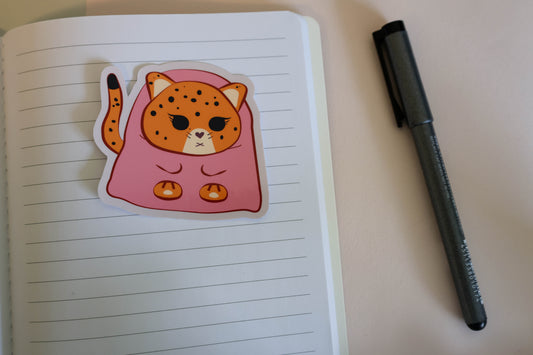 A cute die-cut sticker of a sleepy orange jaguar wrapped in a pink blanket, placed on an open lined notebook next to a black pen.