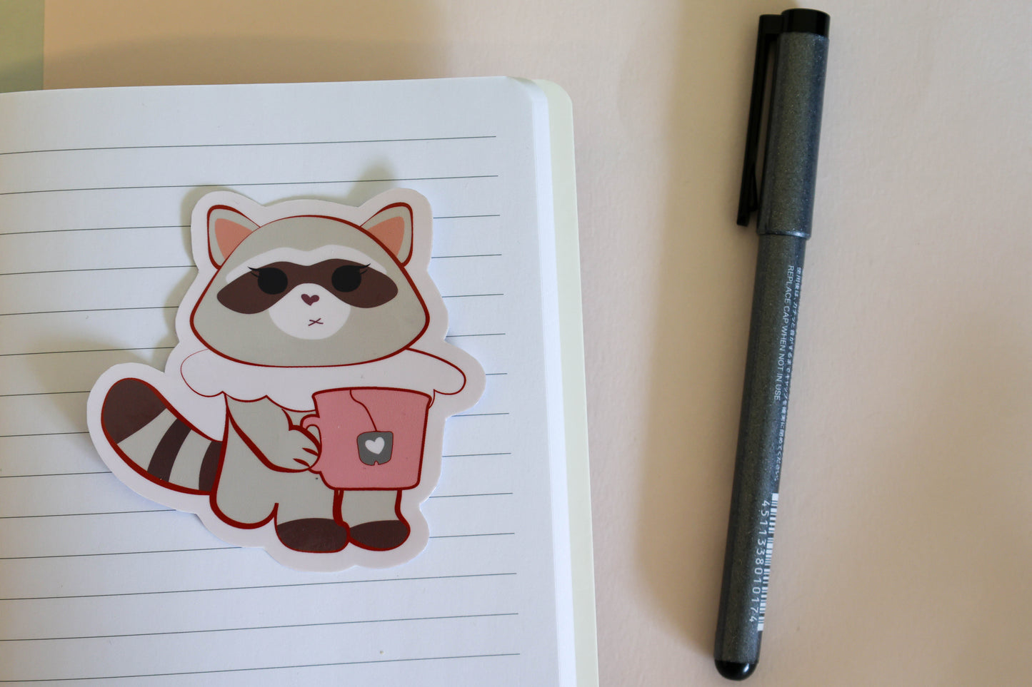 A cute cartoon raccoon sticker and holding a pink mug, placed on a lined notebook next to a black pen.