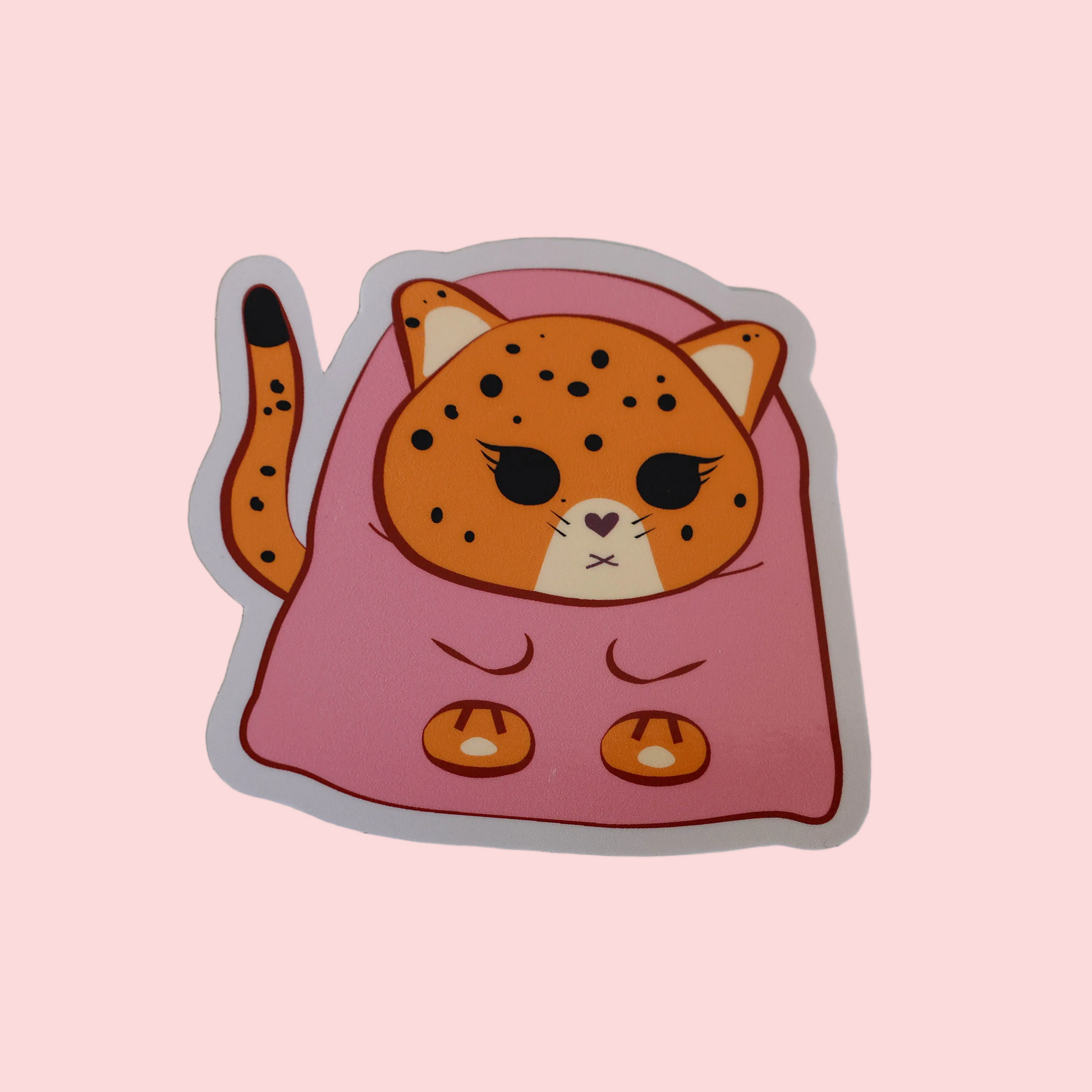 A die-cut sticker featuring a cute cartoon jaguar wrapped in a pink blanket, shown on a solid pale pink background.
