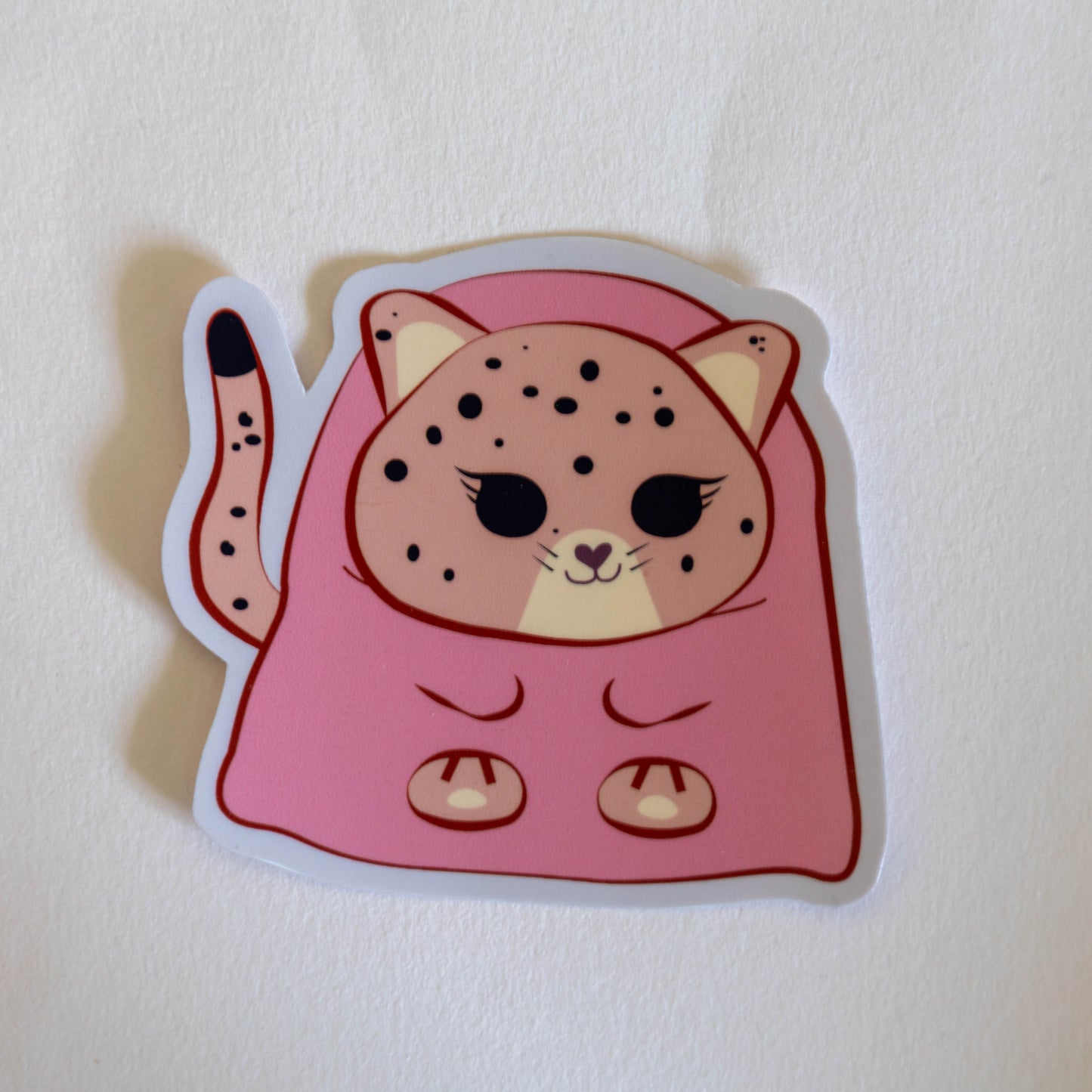 A die-cut sticker featuring a cute cartoon jaguar wrapped in a pink blanket, placed on a textured off-white surface. Cozy Pink Jaguar vinyl sticker for journaling