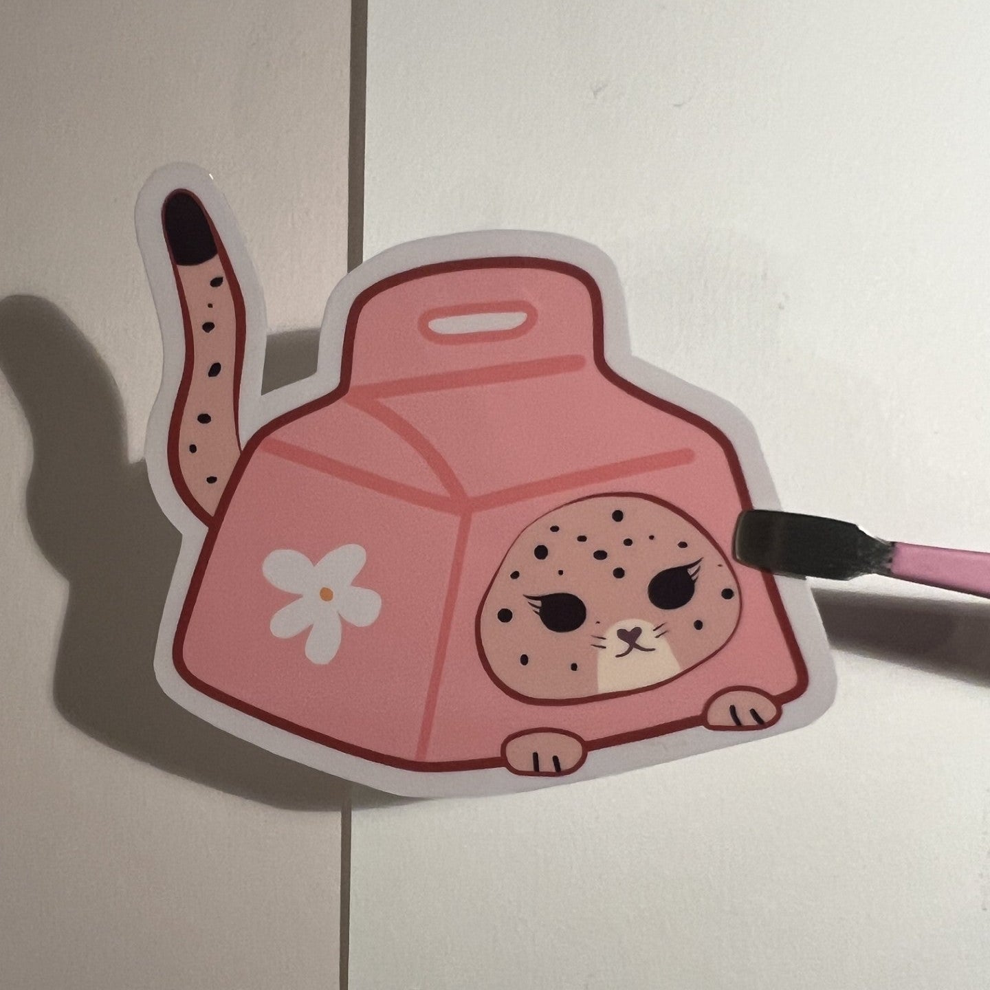 A pink cat inside a milk box sticker or decal with a white flower on the side, shown against a light background with a paintbrush touching the right edge.

