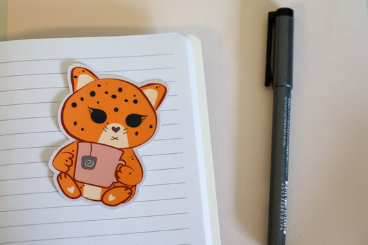 A cute sticker of an orange jaguar cat holding a pink mug, placed on a lined notebook page next to a black pen.
