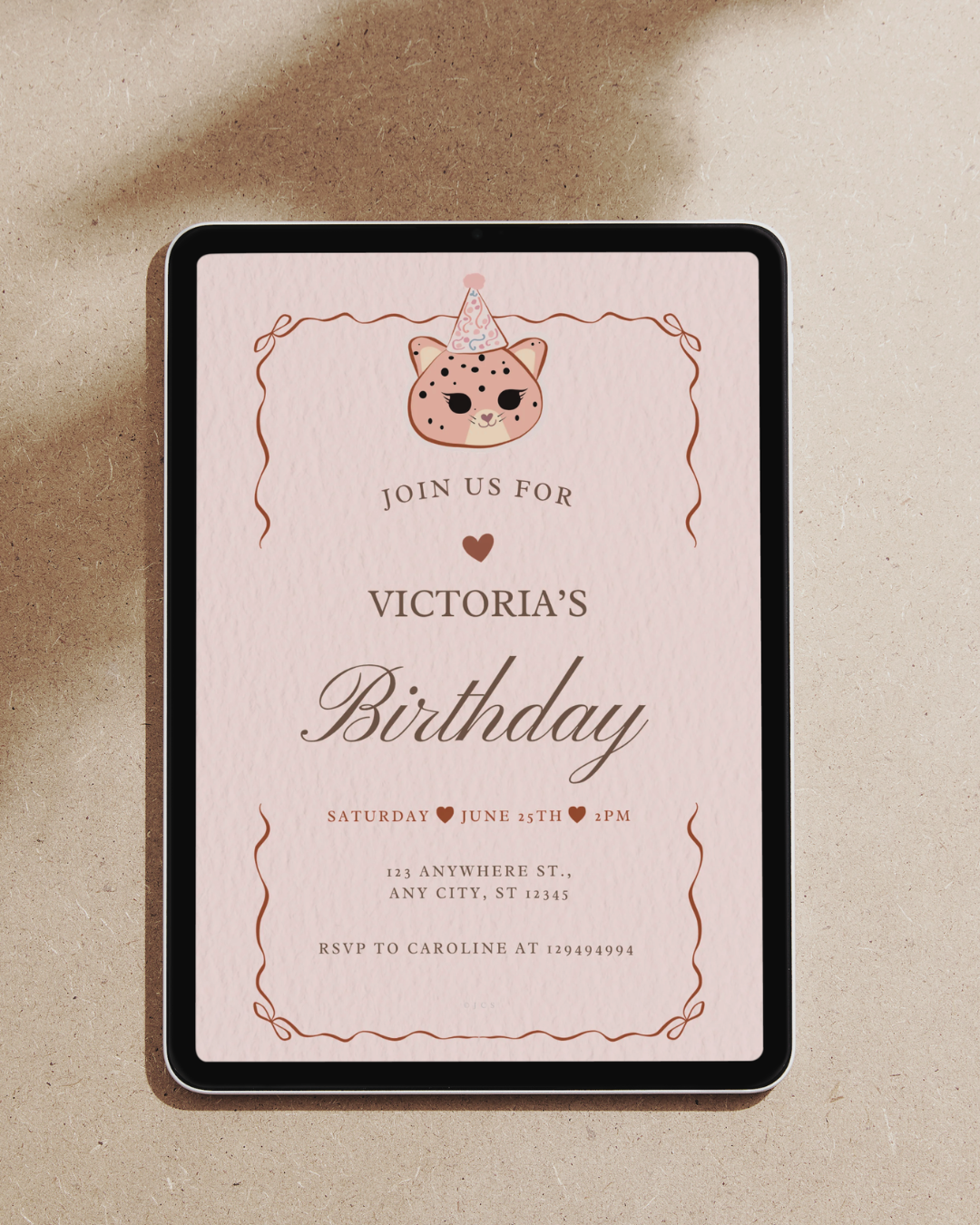 A kawaii birthday invitation card, shown from above on a warm beige textured surface. The card has a pale pink background with a cute cat illustration wearing a party hat at the top, decorative copper-toned border lines, and the text 'JOIN US FOR VICTORIA'S Birthday' with event details below.