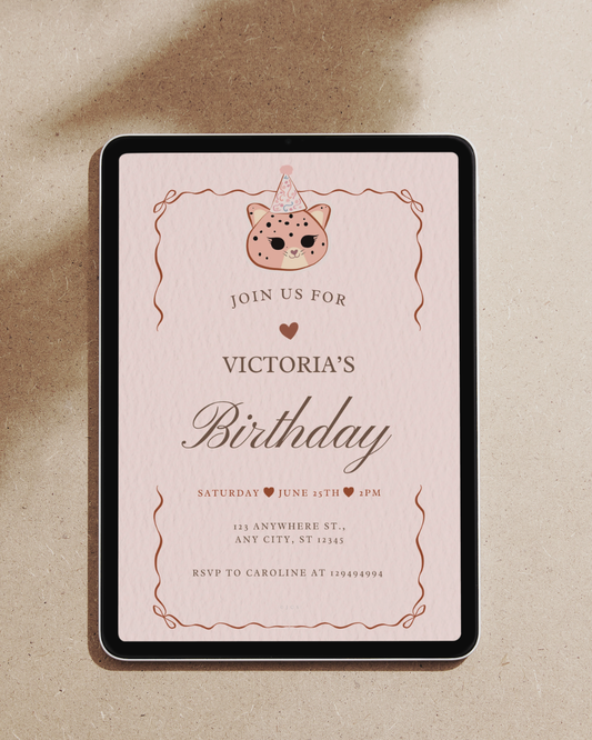 A kawaii birthday invitation card, shown from above on a warm beige textured surface. The card has a pale pink background with a cute cat illustration wearing a party hat at the top, decorative copper-toned border lines, and the text 'JOIN US FOR VICTORIA'S Birthday' with event details below.