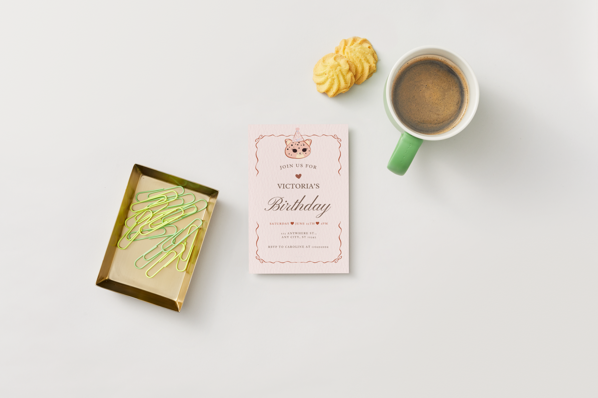 A styled flat-lay featuring a pink and gold birthday invitation card with a kawaii jaguar cat with party hat, accompanied by a green coffee mug, a yellow pastry, and a gold tray with green paperclips on a white background.