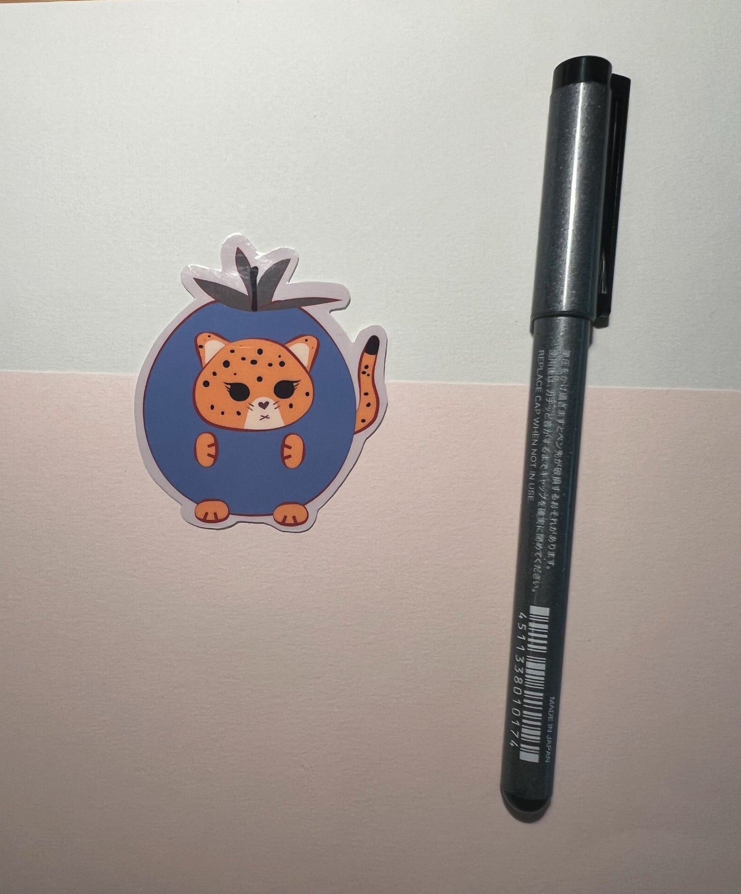A kawaii orange jaguar  inside a blueberry outfit die-cut sticker, and Black pen on a beige surface.