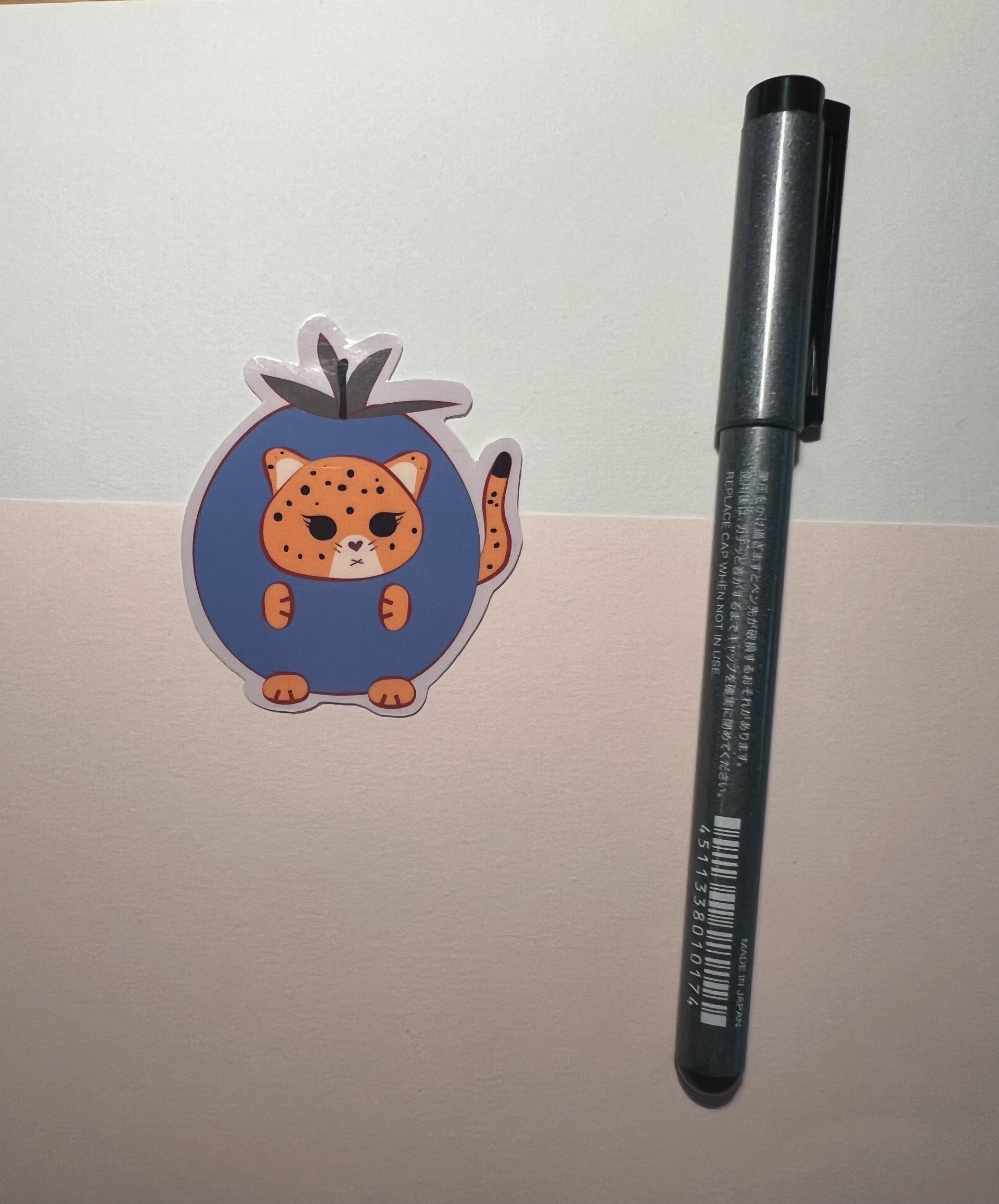 A kawaii orange jaguar  inside a blueberry outfit die-cut sticker, and Black pen on a beige surface.