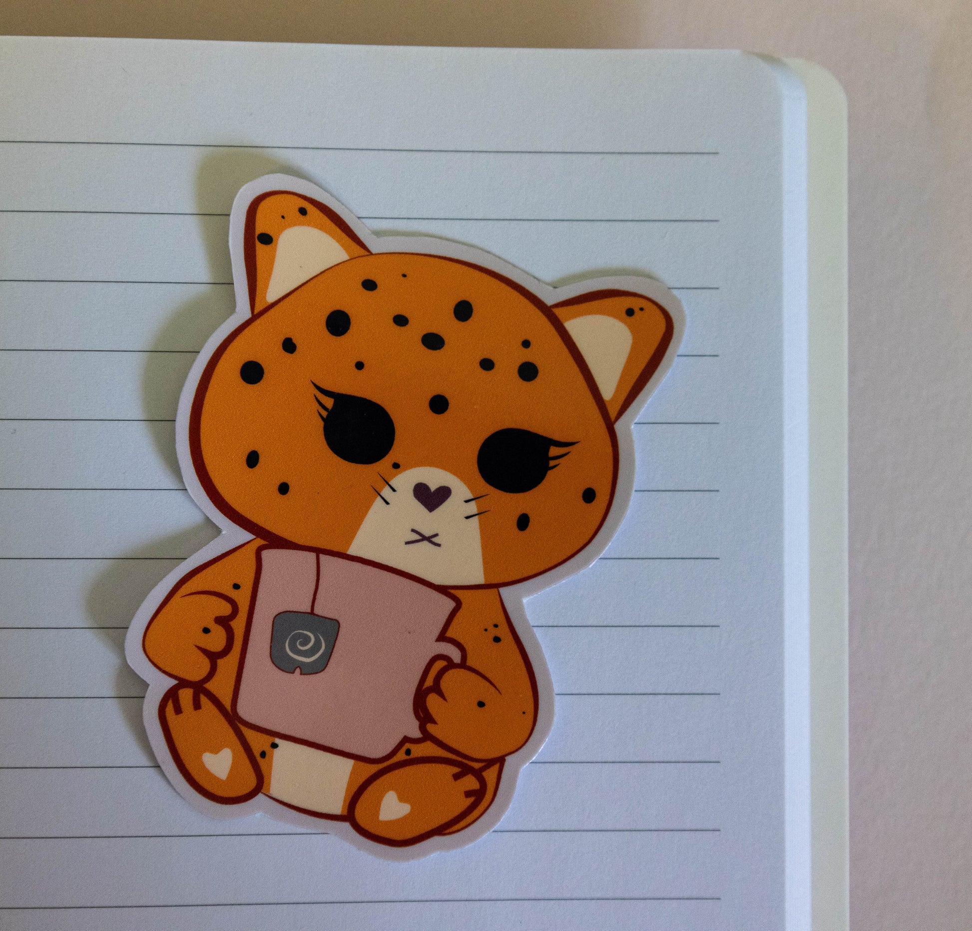 A die-cut sticker of a cute orange jaguar character holding a pink mug, placed on a lined notebook page.