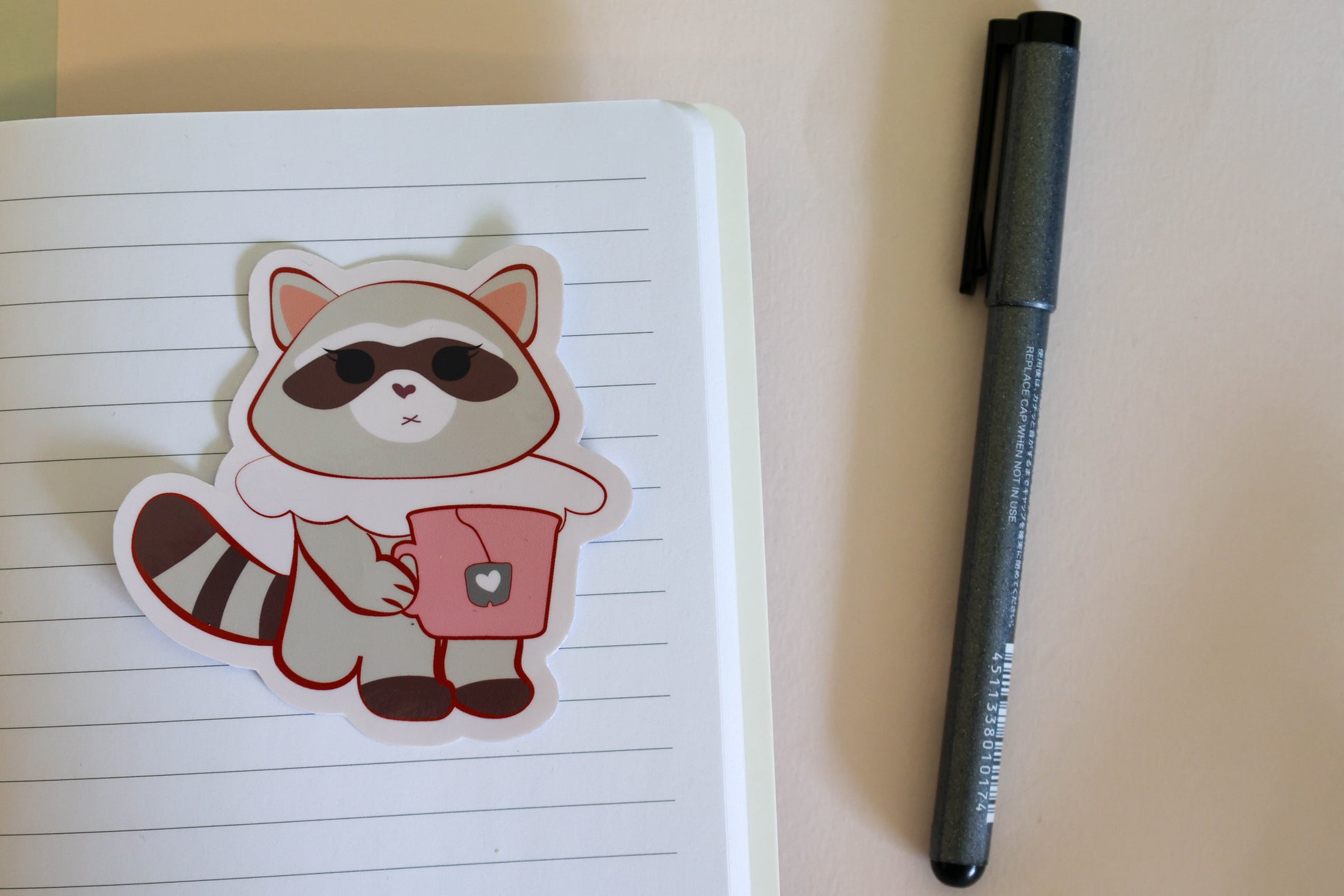 A cute cartoon raccoon sticker  and holding a pink mug, placed on a lined notebook next to a black pen.