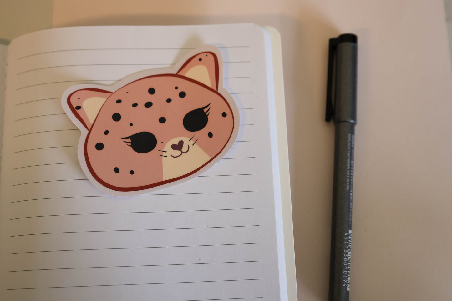 A cute pink die-cut sticker shaped like a jaguar face with black whiskers and spots, placed on an open lined notebook next to a black pen.