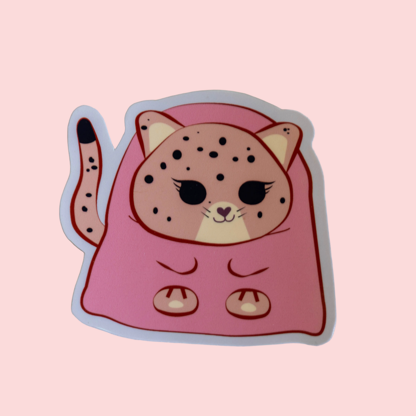 A die-cut sticker featuring a cute cartoon jaguar cat peeking out from a pink blanket, shown on a solid pale pink background.