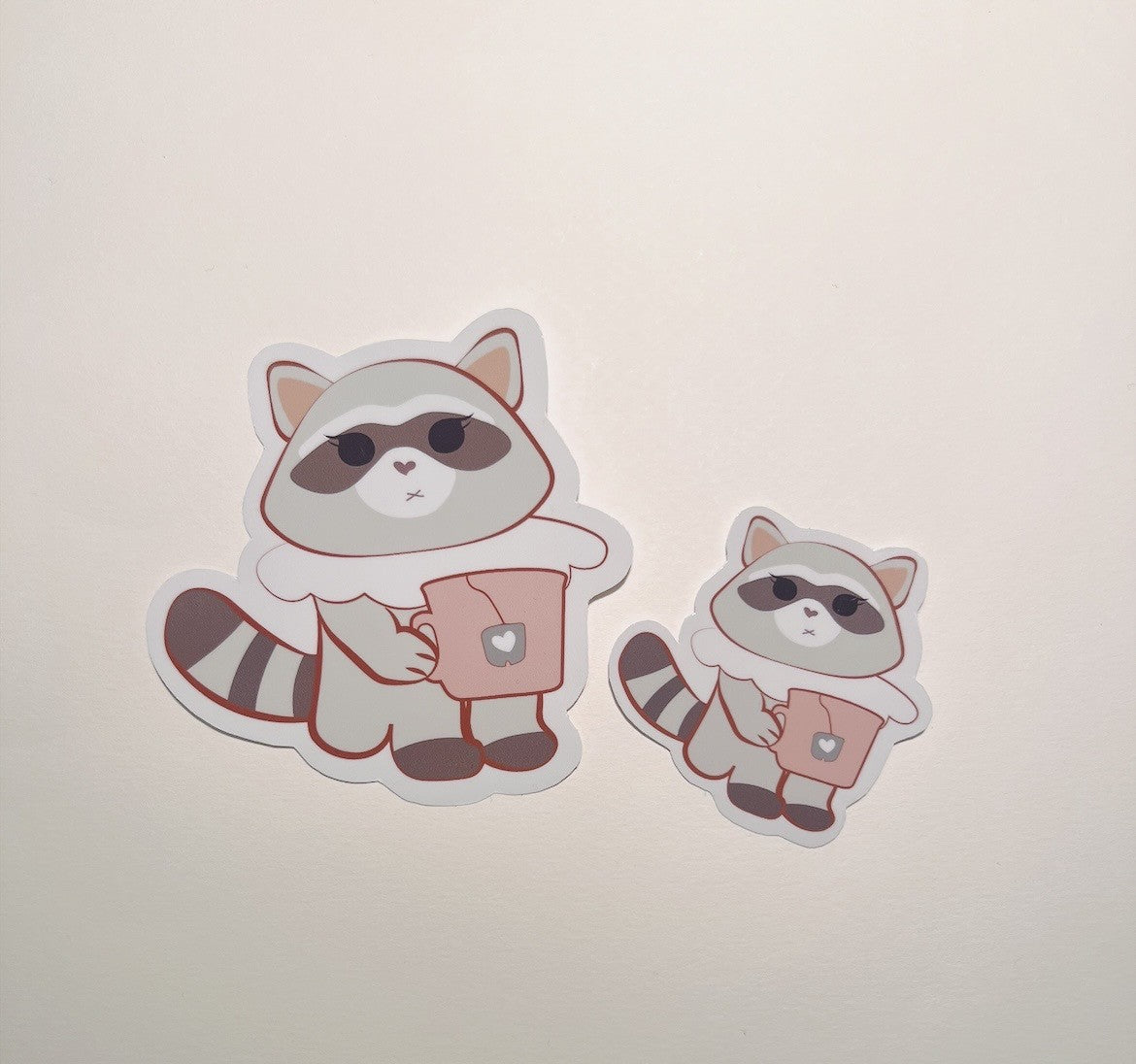 Two raccoon stickers holding a mug, funny stickers,  on a light beige background